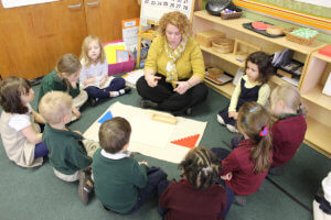 A class of young students and their teacher sitting in a circle around a puzzle
