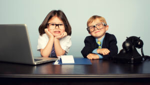 Two young kids sitting at a desk with a phone and a computer
