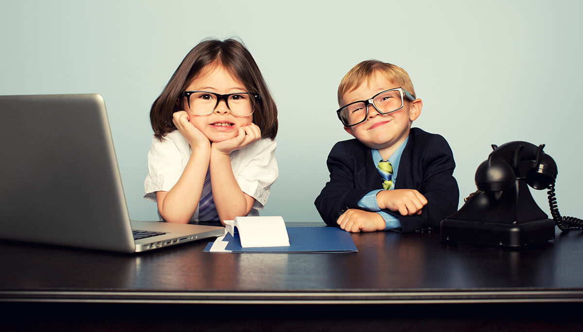 Two young kids sitting at a desk with a phone and a computer